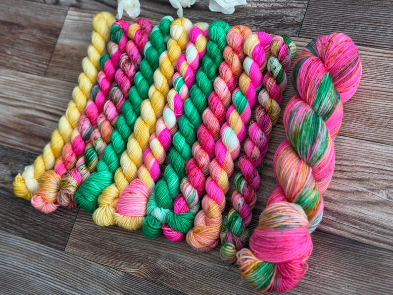 May include: A collection of ten twisted skeins of yarn arranged in a row. The yarn features a vibrant color palette including bright yellow, hot pink, emerald green, and orange, with some skeins displaying variegated patterns of these mixed colors.