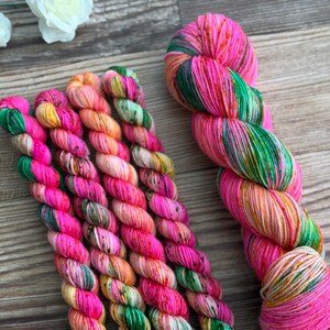 May include: A collection of hand-dyed yarn skeins featuring a vibrant variegated color palette of hot pink, bright green, and soft orange. The yarn is twisted into multiple small bundles and one larger skein, showcasing a speckled texture throughout.