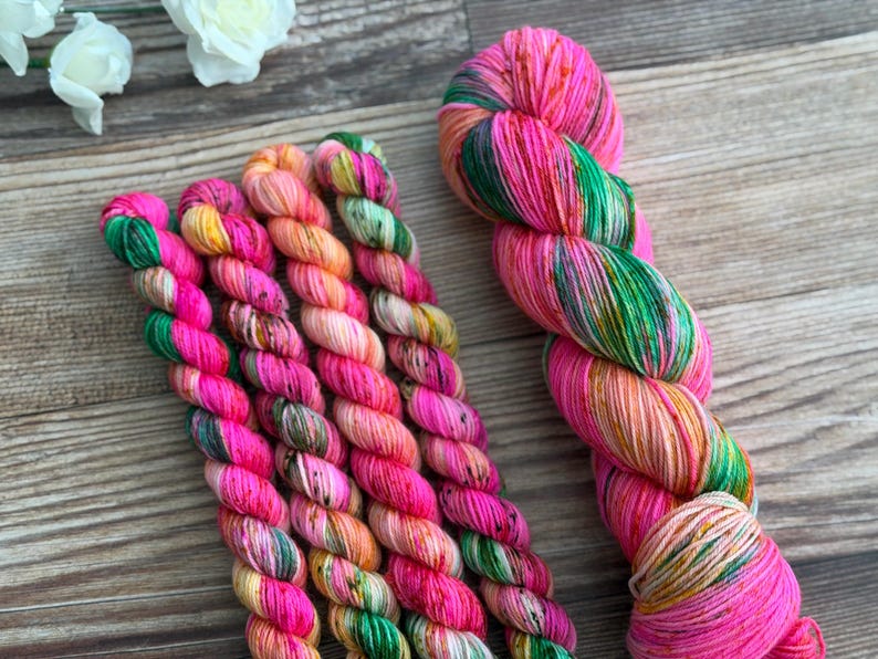 May include: A collection of hand-dyed yarn skeins featuring a vibrant variegated color palette of hot pink, bright green, and soft orange. The yarn is twisted into multiple small bundles and one larger skein, showcasing a speckled texture throughout.