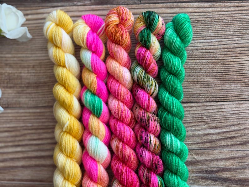 May include: Five twisted skeins of yarn arranged side by side. The first is solid yellow, the second features alternating stripes of neon pink and light green, the third is a variegated orange and pink, the fourth is a mix of green and pink, and the fifth is solid bright green.