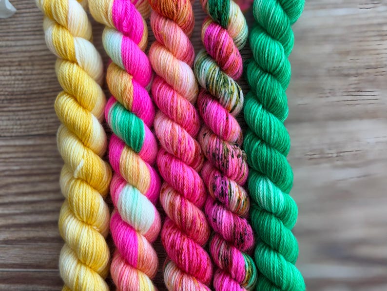 May include: Five twisted skeins of yarn arranged vertically. The first is solid yellow, the second features alternating stripes of neon pink and green, the third is a variegated peach and pink, the fourth is pink with dark speckles, and the fifth is solid green.