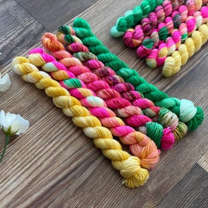 May include: A collection of small, twisted skeins of yarn arranged in a diagonal pattern. The yarn colors include bright yellow, vibrant pink, orange, and green, with some skeins featuring variegated patterns. Three white artificial flowers are visible.