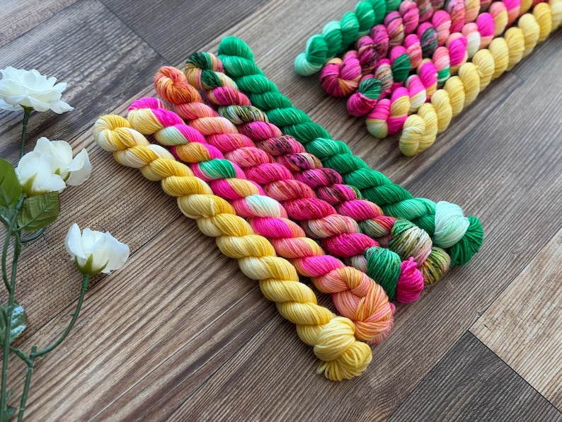 May include: A collection of small, twisted skeins of yarn arranged in a diagonal pattern. The yarn colors include bright yellow, vibrant pink, orange, and green, with some skeins featuring variegated patterns. Three white artificial flowers are visible.
