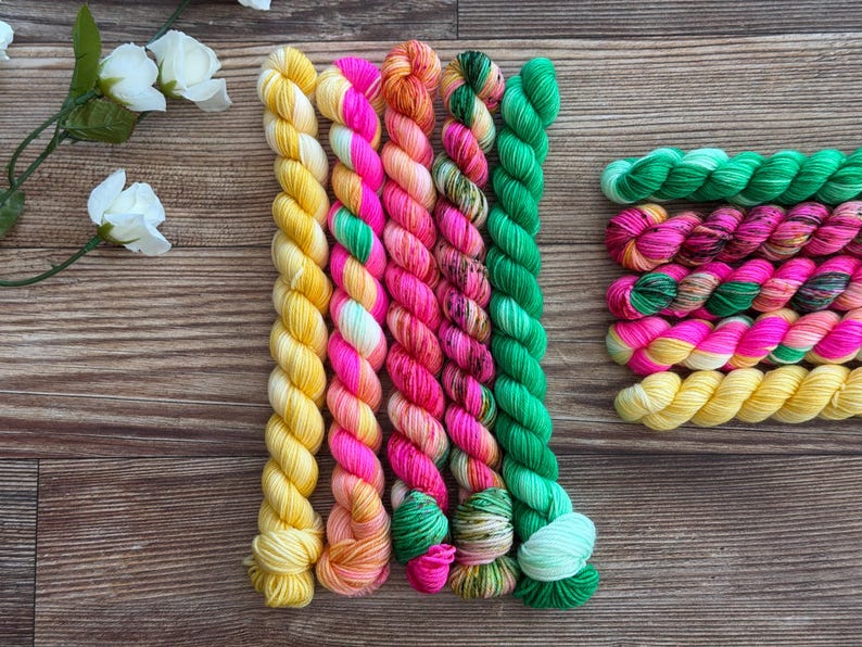 May include: A collection of ten yarn skeins in vibrant colors. Five skeins are arranged vertically, featuring solid yellow, pink and orange variegated, solid pink, multi-colored speckled, and solid green. Five smaller skeins are stacked horizontally.