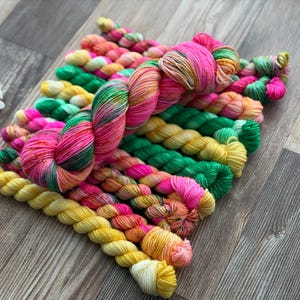 May include: A collection of hand-dyed yarn skeins arranged in a cluster. The yarn features a vibrant color palette including bright pink, sunny yellow, emerald green, and orange, with some skeins displaying variegated patterns of these mixed hues.