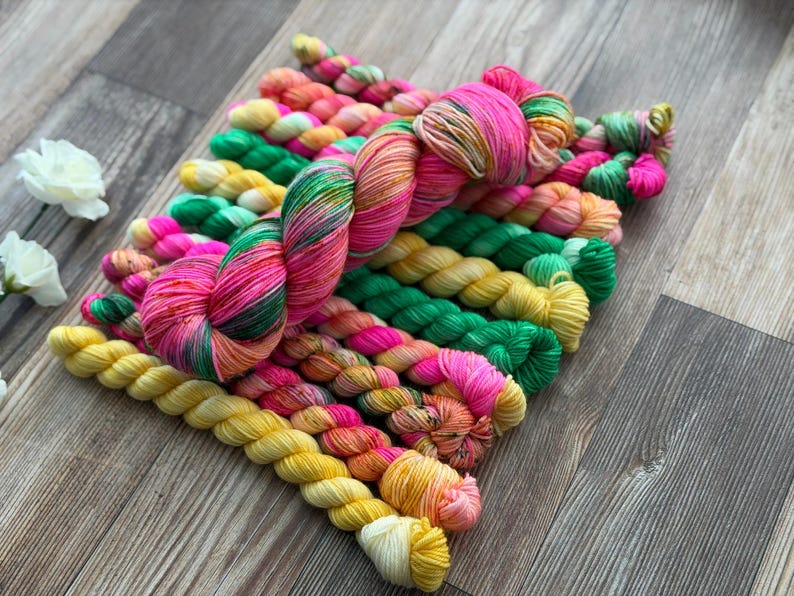 May include: A collection of hand-dyed yarn skeins arranged in a cluster. The yarn features a vibrant color palette including bright pink, sunny yellow, emerald green, and orange, with some skeins displaying variegated patterns of these mixed hues.