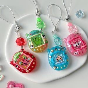 May include: Four colorful digital pet keychains with silver lanyards and decorative beads. The devices are red, yellow-green, blue, and pink, each featuring a unique character on the screen and small printed icons on the plastic casing.
