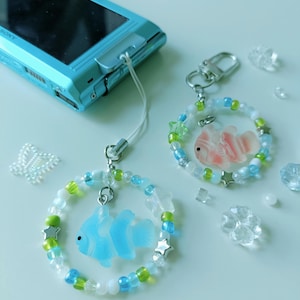 May include: Two beaded fish charms with a blue digital camera. The charms feature a fish pendant in blue and pink, surrounded by a circle of beads in white, blue, and green. Silver star-shaped beads are also included.
