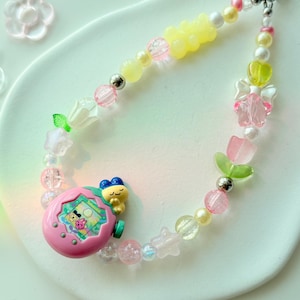 K&ouml;nnte beinhalten: Ein farbenfrohes Perlenarmband mit einem rosa Tamagotchi-Anh&auml;nger, gelben Gummib&auml;rchen-Perlen und verschiedenen pastellfarbenen Perlen. Das Armband enth&auml;lt rosa, wei&szlig;e und transparente Perlen mit silbernen Akzenten. Der Anh&auml;nger hat einen kleinen Bildschirm mit einer Cartoon-Figur.