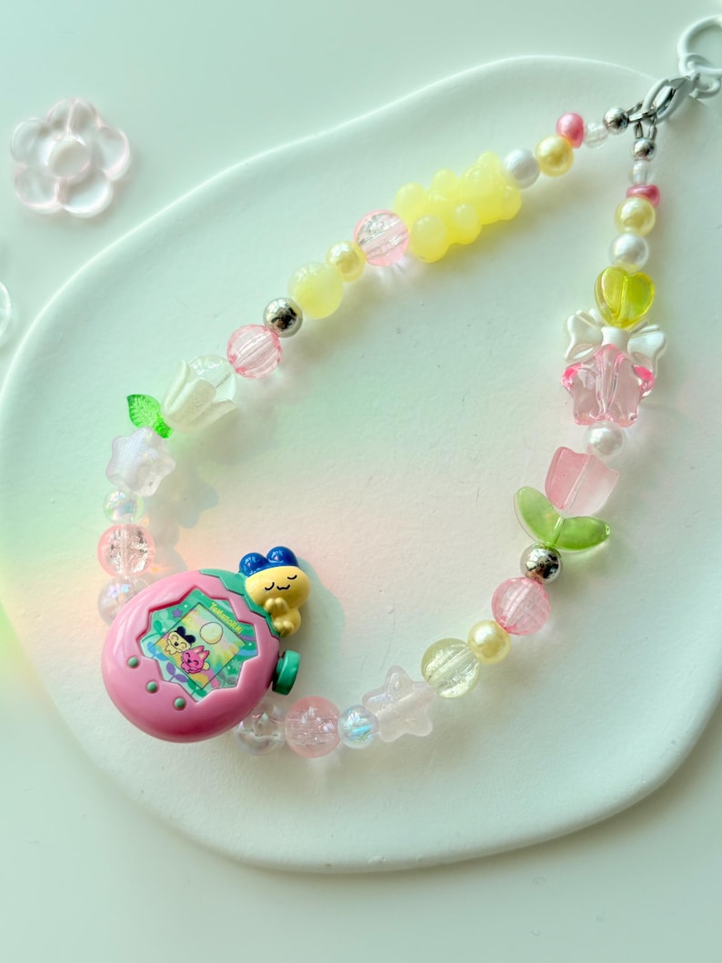 K&ouml;nnte beinhalten: Ein farbenfrohes Perlenarmband mit einem rosa Tamagotchi-Anh&auml;nger, gelben Gummib&auml;rchen-Perlen und verschiedenen pastellfarbenen Perlen. Das Armband enth&auml;lt rosa, wei&szlig;e und transparente Perlen mit silbernen Akzenten. Der Anh&auml;nger hat einen kleinen Bildschirm mit einer Cartoon-Figur.