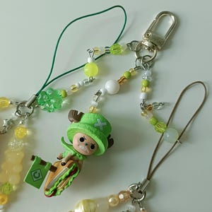 May include: Beaded phone charms in yellow, green, and white. One charm features a cartoon character figure holding a sandwich and a flag. The charms have silver-coloured metal clasps and connectors.