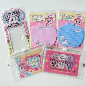 May include: A collection of Esther Bunny stationery items, including a pink heart-shaped photo frame, apple-shaped notepads, and zippered pouches. The items feature cartoon bunny illustrations in pink, blue, and yellow, with the text "ESTHER BUNNY".