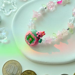 K&ouml;nnte beinhalten: Eine verspielte Halskette mit einem rosa-gr&uuml;nen Dinosaurier-Anh&auml;nger, verziert mit rosa Bl&uuml;ten, transparenten Perlen und kleinen gr&uuml;nen Bl&auml;ttern. Die Halskette ist mit klaren und rosa bl&uuml;tenf&ouml;rmigen Perlen verziert. Japanische M&uuml;nzen und andere W&auml;hrungen sind im Vordergrund zu sehen.