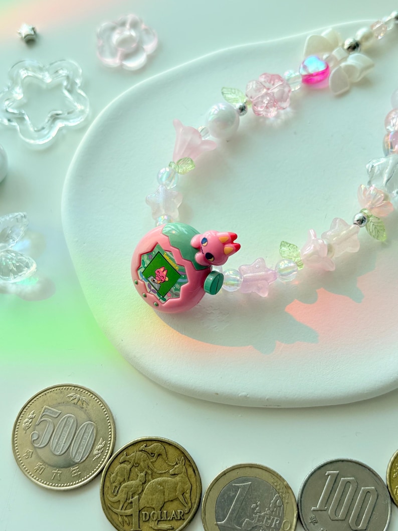 K&ouml;nnte beinhalten: Eine verspielte Halskette mit einem rosa-gr&uuml;nen Dinosaurier-Anh&auml;nger, verziert mit rosa Bl&uuml;ten, transparenten Perlen und kleinen gr&uuml;nen Bl&auml;ttern. Die Halskette ist mit klaren und rosa bl&uuml;tenf&ouml;rmigen Perlen verziert. Japanische M&uuml;nzen und andere W&auml;hrungen sind im Vordergrund zu sehen.