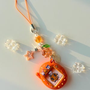 Puede incluir: Un llavero de mascota digital naranja con una pantalla que muestra un personaje de dibujos animados con un gorro de chef. El dispositivo est&aacute; unido a una correa de cuentas con cuentas naranjas en forma de coraz&oacute;n, estrella y flor, y hojas.