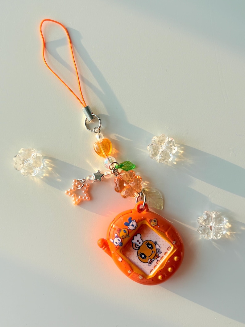 Puede incluir: Un llavero de mascota digital naranja con una pantalla que muestra un personaje de dibujos animados con un gorro de chef. El dispositivo est&aacute; unido a una correa de cuentas con cuentas naranjas en forma de coraz&oacute;n, estrella y flor, y hojas.