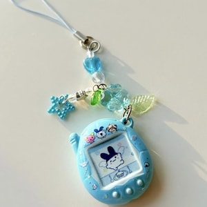 Puede incluir: Un llavero de mascota digital azul claro con una pantalla que muestra un personaje de dibujos animados. El dispositivo est&aacute; unido a una correa de cuentas con un dije de estrella azul, una hoja verde transl&uacute;cida y una cuenta azul en forma de flor.