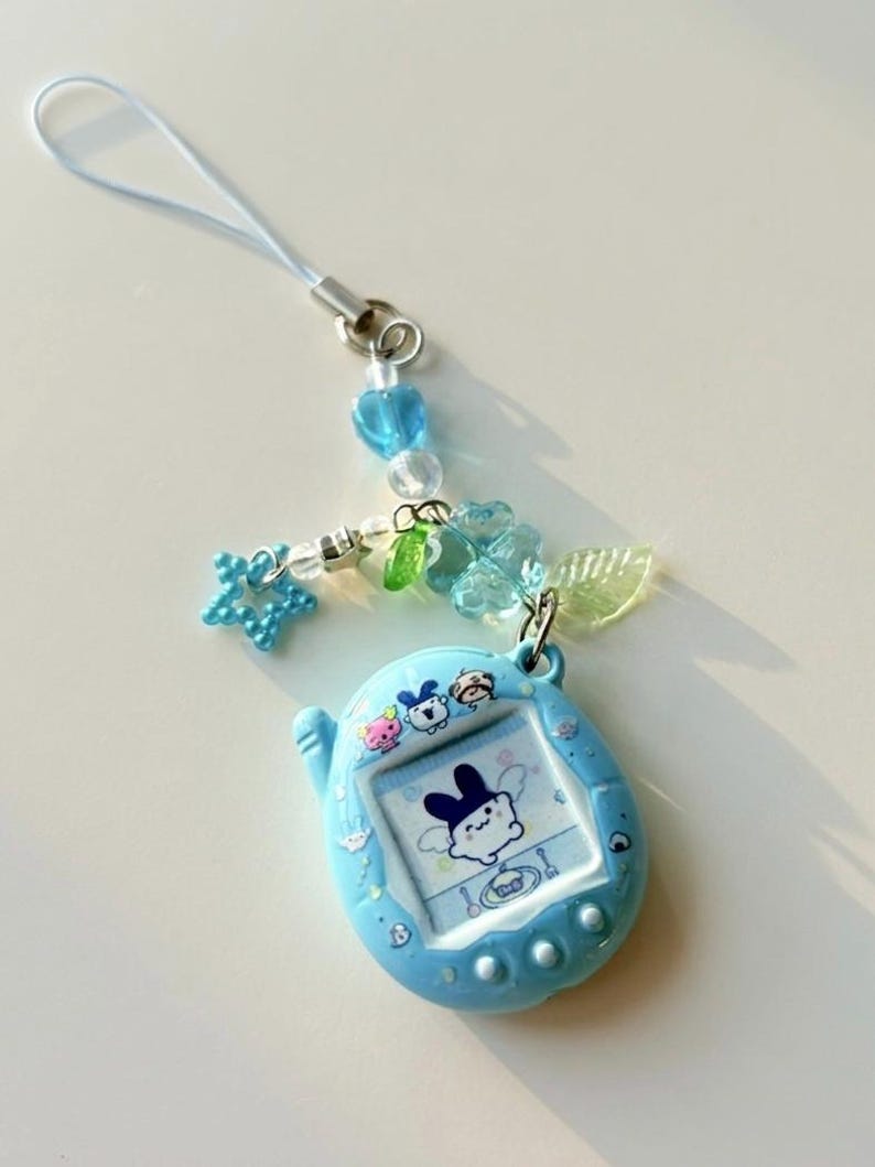 Puede incluir: Un llavero de mascota digital azul claro con una pantalla que muestra un personaje de dibujos animados. El dispositivo est&aacute; unido a una correa de cuentas con un dije de estrella azul, una hoja verde transl&uacute;cida y una cuenta azul en forma de flor.