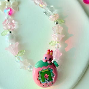 K&ouml;nnte beinhalten: Eine Halskette im pink-gr&uuml;nen Tamagotchi-Design mit Perlen. Die Halskette hat einen pinkfarbenen Tamagotchi-Anh&auml;nger mit einem Hasencharakter, umgeben von pinkfarbenen, transparenten und irisierenden Perlen, Blumen und Schleifen.
