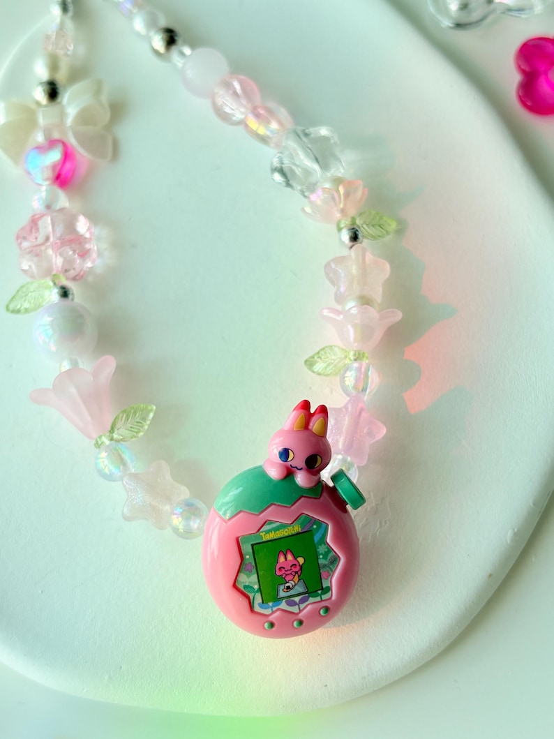 K&ouml;nnte beinhalten: Eine Halskette im pink-gr&uuml;nen Tamagotchi-Design mit Perlen. Die Halskette hat einen pinkfarbenen Tamagotchi-Anh&auml;nger mit einem Hasencharakter, umgeben von pinkfarbenen, transparenten und irisierenden Perlen, Blumen und Schleifen.