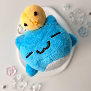 May include: A plush keychain featuring a blue creature with a smiling face and a yellow chick on top. The blue creature has black stitched eyes and mouth. The chick has black eyes and an orange beak. The keychain is on a white surface.