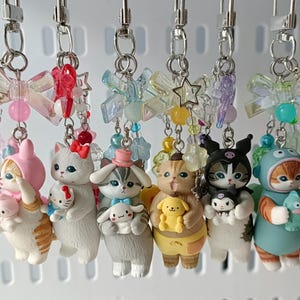 May include: Seven cat-themed keyrings, each with a silver clasp and decorative beads. The figurines depict cats in various outfits, holding small accessories. The keyrings feature a range of colours and designs, perfect for attaching to keys or bags.