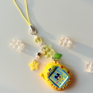 Puede incluir: Un llavero de mascota virtual amarillo con una pantalla que muestra un personaje de dibujos animados, unido a un cord&oacute;n amarillo. El accesorio incluye cuentas decorativas en forma de corazones, estrellas y hojas, junto a flores transparentes.