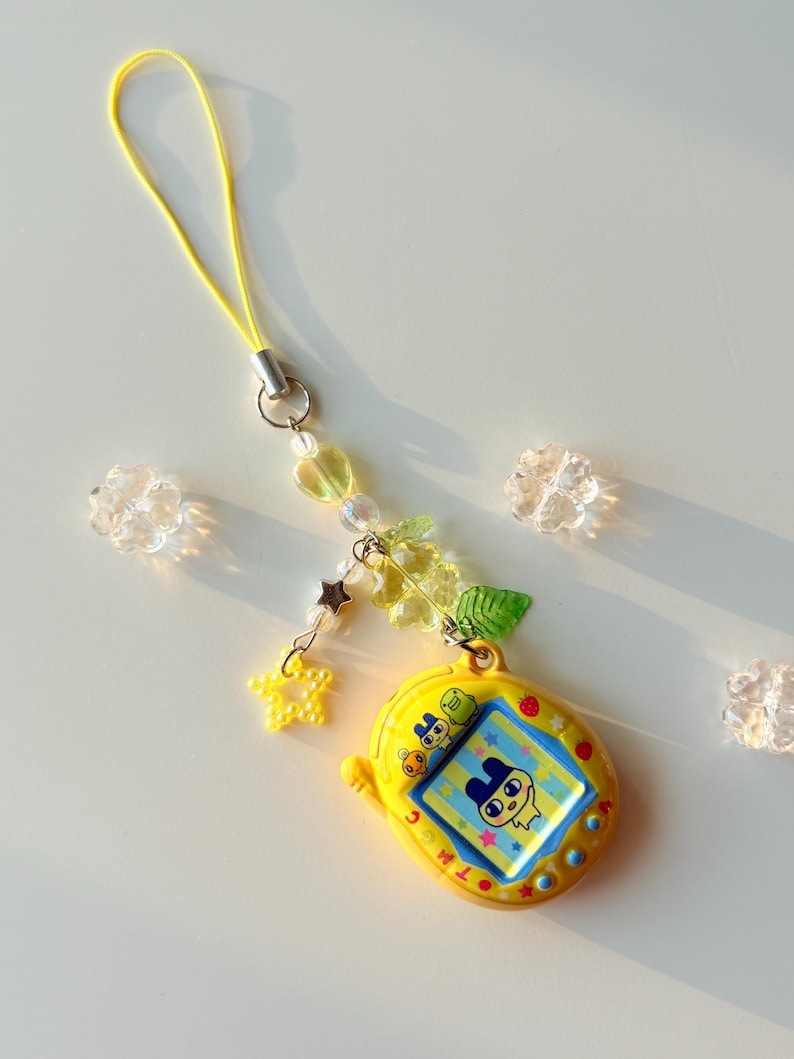 Puede incluir: Un llavero de mascota virtual amarillo con una pantalla que muestra un personaje de dibujos animados, unido a un cord&oacute;n amarillo. El accesorio incluye cuentas decorativas en forma de corazones, estrellas y hojas, junto a flores transparentes.