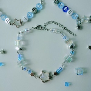 Op de afbeelding: Verschillende handgemaakte armbanden met een hemels thema. De armbanden hebben een mix van heldere, blauwe en witte kralen, zilverkleurige sterbedels en schakels. Sommige armbanden spellen woorden met letterkralen.