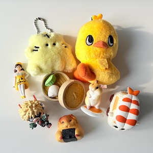 May include: A selection of small, colourful figurines and plush toys. Includes a yellow chick plush, a yellow cat plush with a keychain, a miniature doll, a hamster on a surfboard, a hamster with sushi, a dumpling, and a shrimp sushi.