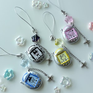 May include: Four silver-toned digital pet charms with screen displays showing characters and the word START. Each charm features a silver cord, a star-shaped bead, and a dangling star charm. Scattered around are small, iridescent, flower-shaped beads.