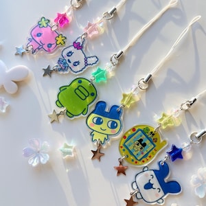 Op de afbeelding: Zes telefoonhangers met kleurrijke acryl figuurhangers, waaronder een roze wezen, een wit konijn, een groene blob, een figuur met blauwe oren, een geel digitaal huisdierapparaat en een blauw-wit gezicht, allemaal versierd met sterrenkralen.
