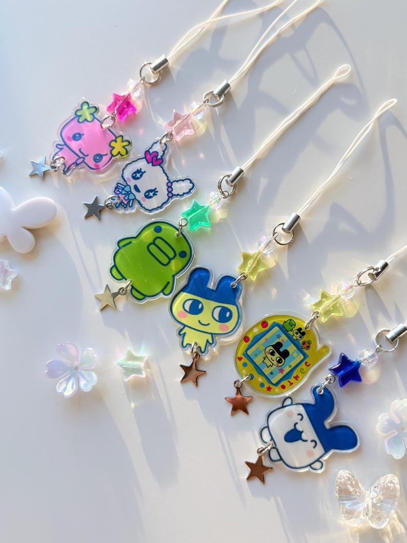 Op de afbeelding: Zes telefoonhangers met kleurrijke acryl figuurhangers, waaronder een roze wezen, een wit konijn, een groene blob, een figuur met blauwe oren, een geel digitaal huisdierapparaat en een blauw-wit gezicht, allemaal versierd met sterrenkralen.