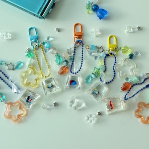 May include: A collection of keychains with colorful charms. Each keychain features a different colored clasp, a chain, and various decorative elements, including star and flower shapes, beads, and small fish-filled cubes. The color palette includes orange, yellow, blue, and green.