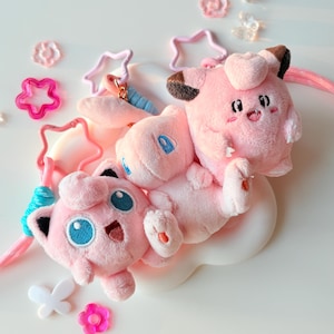 May include: Pink plush toys of Pokémon characters Jigglypuff and Mew, along with star and flower-shaped accessories. The toys have blue eyes and are set against a white background. The accessories are in shades of pink and white.
