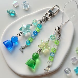 Puede incluir: Dos colgantes de cuentas para bolso con cuentas translúcidas en forma de estrella, cuentas redondas y colgantes en forma de campana en azul y verde. Un colgante incluye una mariposa plateada, el otro un trébol, con letras que forman TAMAGO.