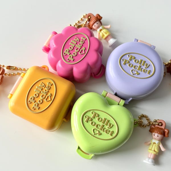 Puede incluir: Cuatro estuches compactos Polly Pocket en rosa, lavanda, amarillo y verde lima, cada uno con una placa ovalada dorada con el texto Polly Pocket y un icono de corazón. Dos pequeñas figuras de muñecas con pelo castaño y trajes amarillos están unidas.