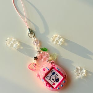 Puede incluir: Un llavero de mascota digital rosa con una pantalla que muestra un conejo blanco. El dije est&aacute; sujeto a un cord&oacute;n rosa y decorado con cuentas transparentes en forma de flor, una cuenta rosa en forma de coraz&oacute;n, una estrella plateada y hojas verdes.