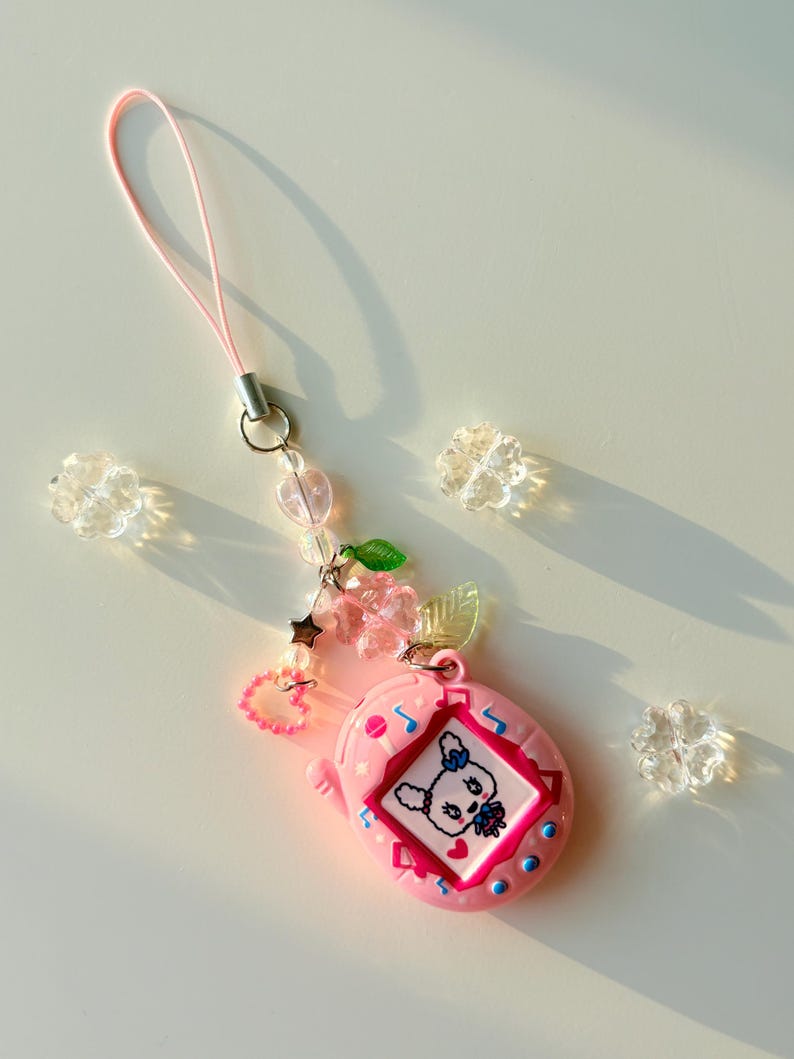 Puede incluir: Un llavero de mascota digital rosa con una pantalla que muestra un conejo blanco. El dije est&aacute; sujeto a un cord&oacute;n rosa y decorado con cuentas transparentes en forma de flor, una cuenta rosa en forma de coraz&oacute;n, una estrella plateada y hojas verdes.