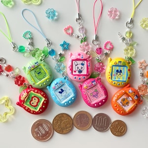 Puede incluir: Una colecci&oacute;n de siete llaveros de mascotas digitales coloridos en rojo, verde, azul, rosa, magenta, amarillo y naranja, cada uno con una pantalla con un personaje, una correa de cuentas decorativa y cinco monedas japonesas para comparar el tama&ntilde;o.