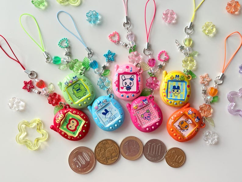 Puede incluir: Una colecci&oacute;n de siete llaveros de mascotas digitales coloridos en rojo, verde, azul, rosa, magenta, amarillo y naranja, cada uno con una pantalla con un personaje, una correa de cuentas decorativa y cinco monedas japonesas para comparar el tama&ntilde;o.