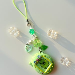 Puede incluir: Un llavero de juguete de mascota digital verde lima con una pantalla que muestra un gr&aacute;fico de criatura verde e iconos de personajes. El dije est&aacute; unido a un cord&oacute;n verde ne&oacute;n con cuentas transparentes y verdes, incluyendo formas de coraz&oacute;n.