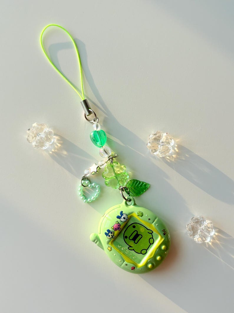 Puede incluir: Un llavero de juguete de mascota digital verde lima con una pantalla que muestra un gr&aacute;fico de criatura verde e iconos de personajes. El dije est&aacute; unido a un cord&oacute;n verde ne&oacute;n con cuentas transparentes y verdes, incluyendo formas de coraz&oacute;n.