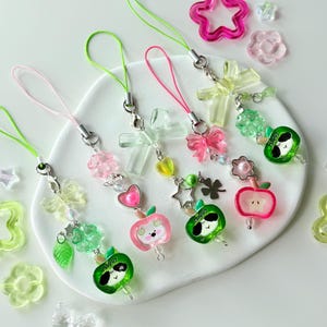 Puede incluir: Una colección de coloridos colgantes de cuentas con varios diseños, incluyendo manzanas, corazones y estrellas. Cada colgante está unido a una cuerda en tonos rosa, verde y amarillo. Formas decorativas adicionales como flores y mariposas están dispersas.