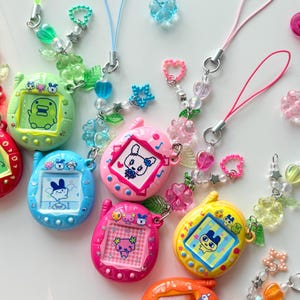 Puede incluir: Una colecci&oacute;n de seis llaveros digitales con forma ovalada en varios colores, incluyendo verde claro, azul, rosa, magenta, amarillo y naranja. Cada dispositivo tiene una pantalla con un personaje y una correa de cuentas con dijes.