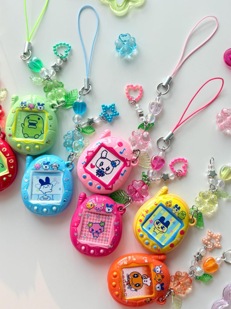 Puede incluir: Una colecci&oacute;n de seis llaveros digitales con forma ovalada en varios colores, incluyendo verde claro, azul, rosa, magenta, amarillo y naranja. Cada dispositivo tiene una pantalla con un personaje y una correa de cuentas con dijes.