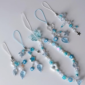 Puede incluir: Varios colgantes de teléfono con cuentas en tonos azules, blancos y transparentes. Los colgantes presentan varias formas, incluyendo estrellas, corazones, flores y peces. Algunos colgantes tienen un acento en forma de mariposa o flor. Cada colgante tiene un lazo blanco o azul.