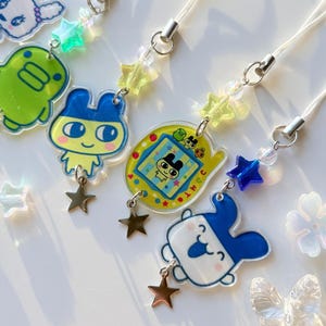 Op de afbeelding: Een collectie telefoonhangers met kleurrijke acryl figuurhangers, waaronder een groen wezen, een personage met blauwe oren en een geel digitaal huisdierapparaat. Elke hanger is versierd met doorzichtige sterrenkralen en een kleine metalen ster.