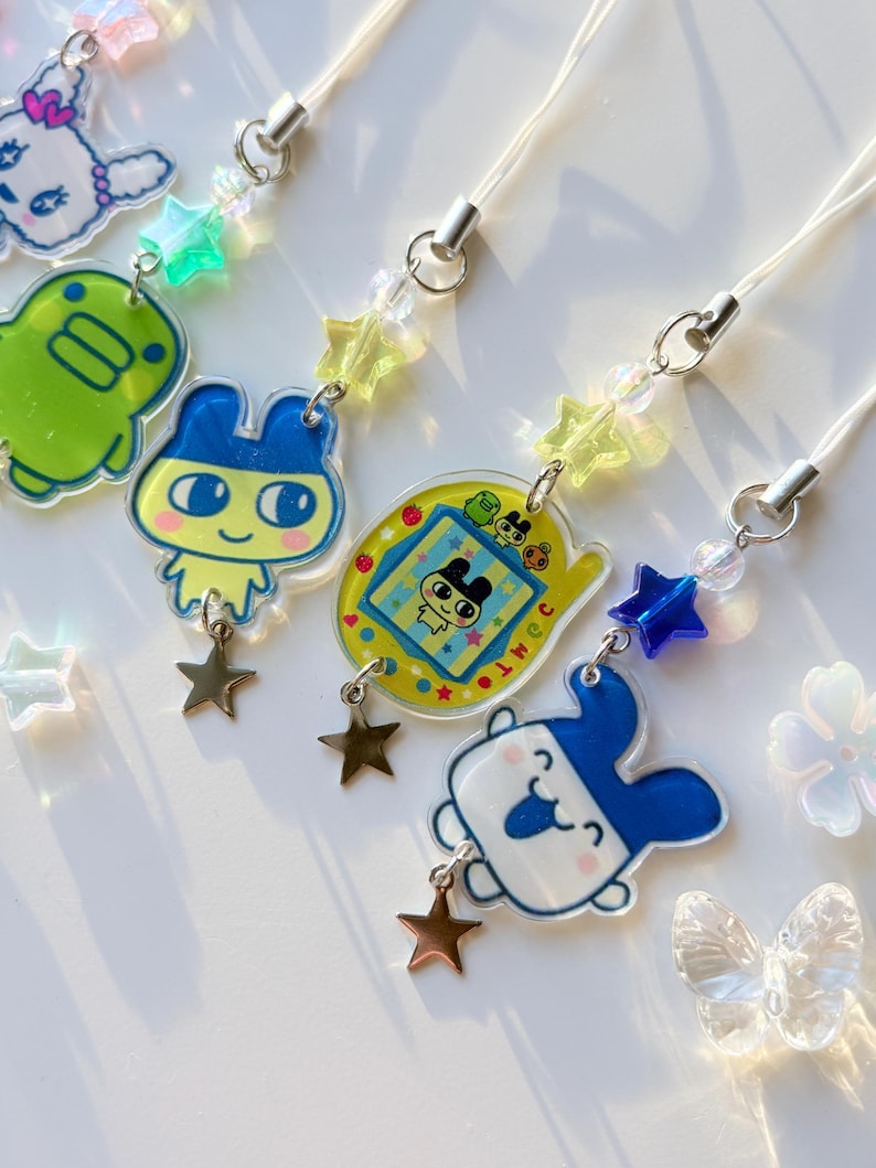 Op de afbeelding: Een collectie telefoonhangers met kleurrijke acryl figuurhangers, waaronder een groen wezen, een personage met blauwe oren en een geel digitaal huisdierapparaat. Elke hanger is versierd met doorzichtige sterrenkralen en een kleine metalen ster.