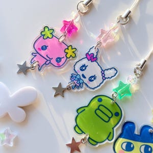 Op de afbeelding: Vier acryl telefoonhangers met kleurrijke stripfiguren, waaronder een roze wezen, een wit konijn, een groene blob en een blauw dier. Elke hanger is bevestigd aan een wit koord met kleine stervormige kralen en een zilverkleurige sterbedel.